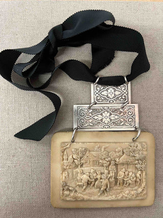 A necklace that is a true work of art. It consists of a bas-relief in meerschaum, depicting a 19th century village feast decorated with two engraved silver-plated rectangles, whose motif is reminiscent of the French gardens designed by architect and draftsman Andrè Le Nôtre. The whole is on a black 'gros-grain' ribbon, which gives balance to the piece.