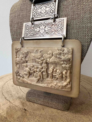A necklace that is a true work of art. It consists of a bas-relief in meerschaum, depicting a 19th century village feast decorated with two engraved silver-plated rectangles, whose motif is reminiscent of the French gardens designed by architect and draftsman Andrè Le Nôtre. The whole is on a black 'gros-grain' ribbon, which gives balance to the piece.