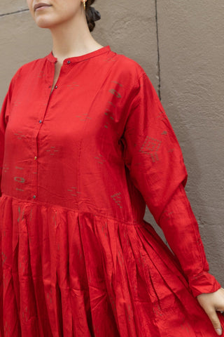 This silk dress in scarlet red from Injiri can be worn from day to night. Cut from silk to an oversized silhouette, it has a round neckline and a row of button till the waist, from there onwards, the silhouette widens thanks to pleats on the dress that give more volume to the lower part. This model features three-quarter length sleeves.