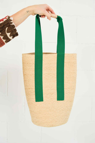 This long and tapered tote is stitched from a 5/16” raffia tape sewn edge to edge for flexibility. A small inside pocket will hold a phone and credit cards. The 1 5/8” emerald green elastic handle straps stops halfway down the bag, and a natural leather handle narrows the strap for a better hold. This bag can be carried over the shoulder, and when loaded, the elastic straps make it bounce along.